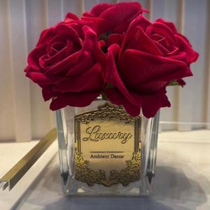May include: A clear glass square vase filled with deep red artificial roses. The front of the vase features a decorative gold-colored label with an ornate border, displaying the text Luxury and Ambient Decor in a black script and serif font.
