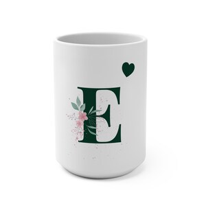 Könnte beinhalten: Weißer Keramikbecher mit einem dunkelgrünen Buchstaben "E", verziert mit rosa Blumen und einem grünen Herz. Der Becher hat eine glänzende Oberfläche. Das Design ist schlicht und elegant, für Heißgetränke.