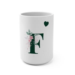 Può includere: Tazza in ceramica bianca con la lettera "F" verde scuro, decorata con fiori rosa e un cuore verde. Il design della tazza è semplice ed elegante, adatta per bevande calde. La tazza è alta circa 10 cm.