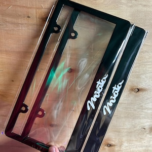 May include: A pair of rectangular black number plate frames encased in clear plastic packaging. Each frame features the word Miata written in a white cursive script along the bottom edge. The frames include pre-drilled mounting holes.