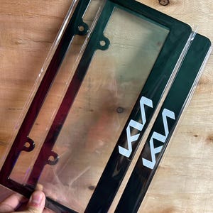 May include: A pair of glossy black license plate frames featuring the white Kia logo printed on the bottom right corner. The frames are rectangular with rounded corners and four mounting holes, encased in clear protective plastic packaging.