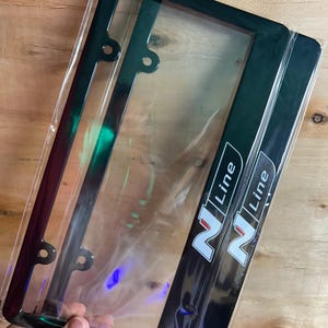 May include: A pair of rectangular black license plate frames featuring the N Line logo in white with a red accent. The frames are encased in clear plastic packaging and include pre-drilled mounting holes along the top and bottom edges.