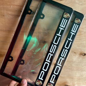 May include: Two black rectangular license plate frames featuring the white text PORSCHE printed vertically along the right side. Each frame includes a small, colorful Porsche crest emblem at the top and bottom corners of the right-hand side.