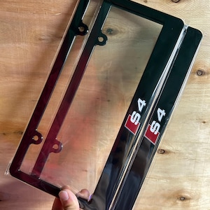 Peut inclure: Deux cadres de plaque d'immatriculation noirs emballés dans du plastique transparent. Chaque cadre présente un emblème rectangulaire rouge avec le texte blanc S4 situé en bas à droite. Les cadres ont quatre trous de montage circulaires.
