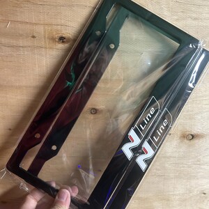 May include: Black license plate frame with the text "N Line" in white. The frame is rectangular with rounded corners and has pre-drilled holes for mounting. The frame is packaged in clear plastic.
