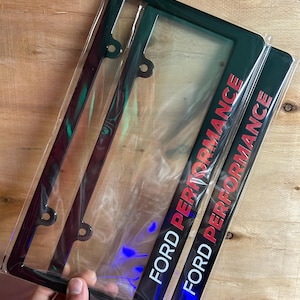 May include: Two black rectangular number plate frames made of a glossy material, each featuring the text FORD PERFORMANCE printed in bold white and red lettering. The frames are encased in clear protective plastic packaging for display.
