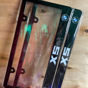 May include: Two black license plate frames for a BMW X5, each featuring the white X5 logo and the blue and white BMW circular emblem. The frames are encased in clear plastic protective packaging and have pre-drilled holes for mounting.