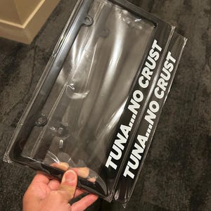 May include: A black rectangular number plate frame encased in clear plastic packaging. The frame features the white text TUNA...NO CRUST printed vertically along the right side. The design includes four mounting holes for attachment to a vehicle.