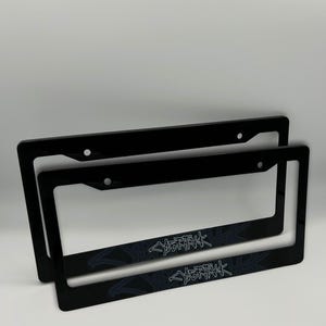 May include: Two black license plate frames with the word "CYBERPUNK" in a stylized font. The frames are rectangular with rounded corners and have pre-drilled holes for mounting. The text is in a light gray color.