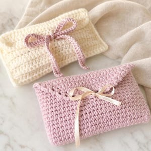 May include: Two rectangular crochet pouches with envelope-style flaps. One pouch is cream-coloured with a matching crocheted tie closure, while the other is dusty rose with a thin, smooth, light-coloured ribbon tied into a bow at the centre.