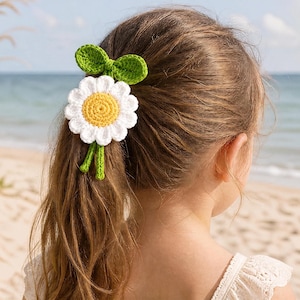 May include: A crochet hair accessory featuring a white daisy flower with a yellow center, accented by two green leaves at the top and two thin green stems hanging down, secured in a ponytail to hold the hair in place with its decorative design.