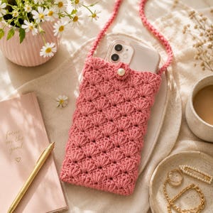May include: A pink crochet phone pouch featuring a textured shell stitch pattern and a single pearl button closure. The pouch includes a matching long crochet strap and holds a smartphone with a visible dual-lens camera system on the back.