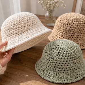 Könnte beinhalten: Drei gehäkelte Fischerhüte in Creme, Beige und Salbeigrün. Jeder Hut hat ein offenes Webmuster an der Krone und eine feste, eng gestrickte Krempe. Die Hüte sind aus Garn gefertigt und haben eine abgerundete Form mit einer kurzen, abfallenden Krempe.