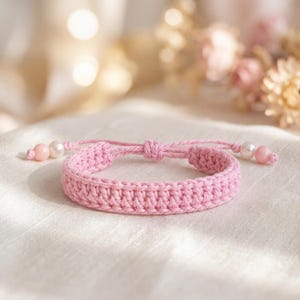 May include: A light pink crocheted bracelet featuring a central adjustable sliding knot. The ends of the cord are decorated with small, round, white pearl-like beads and larger, smooth, light pink spherical beads on each side of the band.