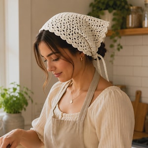 May include: A cream-colored crochet headscarf featuring an intricate openwork pattern and a scalloped edge. The triangular head covering is tied at the back with two thin crochet strings, providing a secure fit while showcasing detailed needlework.
