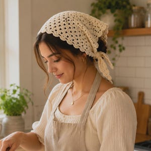 May include: A cream-colored, crocheted headscarf with a scalloped edge and tie closure. The head covering is worn by a person wearing a light-colored apron and top. The kitchen setting includes plants, wooden utensils, and a window.