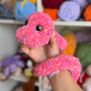 May include: A handmade pink plush snake with black safety eyes and colorful flecks. The soft, cuddly toy is made of a textured yarn and is designed to be a child's toy.