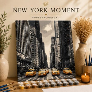 Könnte beinhalten: Ein Malen-nach-Zahlen-Set mit dem Titel NEW YORK MOMENT, das eine schwarz-weiße Leinwand einer Stadtstraße mit vier gelben Taxis zeigt. Das Set enthält nummerierte Farbtöpfe, mehrere Pinsel und eine gedruckte Anleitung für den Malprozess.