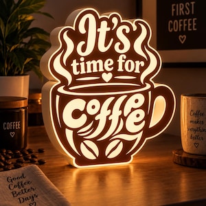 Può includere: Un'insegna luminosa bianca a forma di tazza da caffè con il testo "It's time for coffee" in un carattere stilizzato. L'insegna emette una luce calda, mettendo in risalto il design della tazza, i vortici di vapore e i chicchi di caffè.