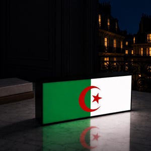 May include: A rectangular light box displaying the Algerian flag, with green and white sections, a red crescent, and a star. The box sits on a marble surface, reflecting the flag's image. The background shows a dark room and a city view.