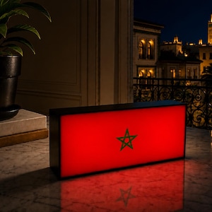 Peut inclure: Un caisson lumineux rectangulaire affichant le drapeau marocain, avec un fond rouge et une étoile verte. Le caisson est posé sur une surface en marbre, avec une vue sur une ville la nuit par une fenêtre.