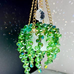 May include: A hanging ornament featuring a small white pot with cascading strands of faceted green crystal beads. The plant is suspended by gold-toned metal chains and includes a single clear, multifaceted crystal prism at the top center.