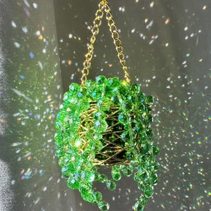 May include: A hanging decorative planter with a gold chain and a wire frame. The planter is filled with green, faceted beads, creating a cascading, plant-like appearance. The background has a soft, shimmering effect.