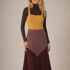 Pode incluir: Uma camisola de gola alta castanha escura de manga comprida usada sob uma regata de malha amarelo-mostarda com decote quadrado e bainha com padrão chevron. O conjunto inclui uma saia midi com design bicolor e um painel chevron malva.