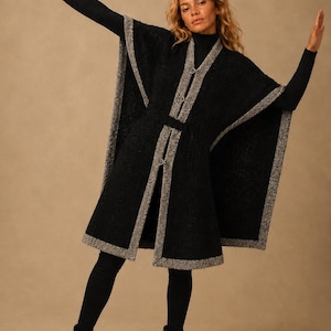 May include: A black sleeveless poncho featuring a textured finish and a contrasting grey speckled trim along the edges. The garment includes a central front closure with two metal clasps and a matching fabric belt at the waist, worn over a black long-sleeved top, black leggings, and black ankle boots.