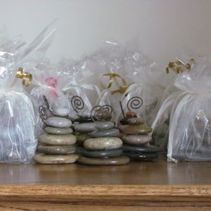 May include: Five small stacks of smooth, gray stones are wrapped in white organza bags with gold ribbon. Each stack is topped with a decorative wire swirl.