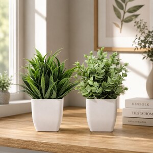 May include: Two artificial potted plants in square white ceramic containers. The left plant features long, pointed, dark green grass-like leaves. The right plant has rounded, light green eucalyptus-style foliage. Both sit on a wooden surface.