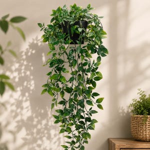 May include: A hanging artificial plant with cascading green leaves in a black plastic pot. The foliage features various shades of green with pointed, oval-shaped leaves that drape downward, creating a dense, trailing appearance from the container.