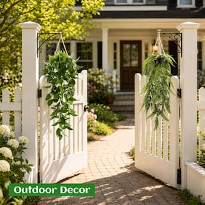 May include: Two hanging artificial plants suspended from black metal wall brackets on a white picket fence gate. One plant features broad, pointed green leaves, while the other is a trailing fern with delicate, feathery green fronds.