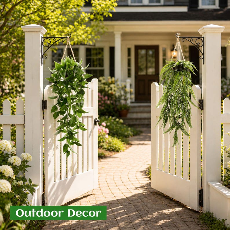 May include: Two hanging artificial plants suspended from black metal wall brackets on a white picket fence gate. One plant features broad, pointed green leaves, while the other is a trailing fern with delicate, feathery green fronds.