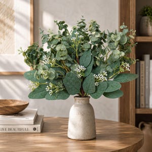 May include: A bouquet of artificial eucalyptus stems with rounded green leaves and small white flower clusters arranged in a light beige, textured ceramic vase. The plant features varied shades of sage and forest green foliage on thin stems.
