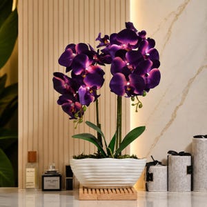 May include: An artificial orchid plant with deep purple and magenta petals, featuring green stems and leaves, potted in a white, horizontally ribbed oval ceramic container that rests on a light-colored wooden base.