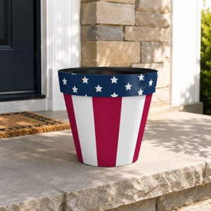 Peut inclure: Un pot de fleurs cylindrique et conique arborant un motif patriotique avec une bande supérieure bleu foncé ornée d'étoiles blanches, et une section inférieure à rayures verticales rouges et blanches alternées. Le bord intérieur est noir.