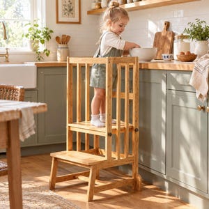 May include: A light-colored wooden learning tower with a step stool base and vertical safety railings. The structure features a platform for standing, side supports, and a lower step, designed to provide elevation for reaching kitchen counters.