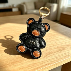 Puede incluir: Un llavero con forma de oso en negro y marrón con acabado texturizado tipo cuero, costuras blancas en contraste y una anilla de metal dorado. El pecho del oso lleva las letras SM en dorado, con detalles en marrón en las orejas, el hocico y los pies.