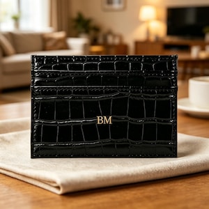 May include: A black card holder with a glossy crocodile-embossed texture. The rectangular accessory features horizontal slots for cards and is personalized with the gold-colored capital letters BM centered on the front surface of the item.