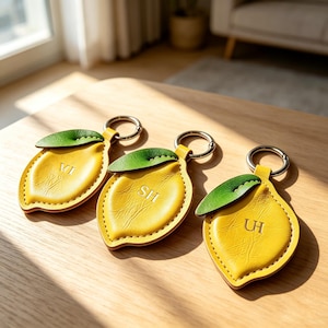 May include: Three lemon-shaped keychains made of yellow textured material with a green leaf accent at the top. Each keychain features a silver metal ring and is embossed with unique two-letter initials: VI, SH, and UH, centered on the lemon body.