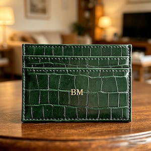 May include: A dark green card holder featuring a textured crocodile skin pattern and visible stitching along the edges. The front of the card holder displays the gold embossed initials BM centered on the surface, with two horizontal card slots visible.