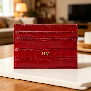 May include: A rectangular red card holder with a glossy crocodile skin texture and visible stitching along the edges. The front features the gold embossed letters BM centred in the middle. It has multiple horizontal slots for storing cards.
