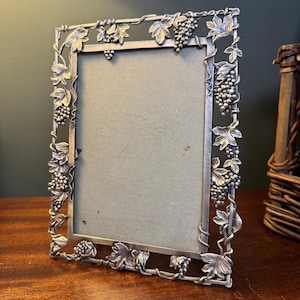 May include: A rectangular silver-toned metal picture frame featuring a decorative border of sculpted grape clusters and vine leaves. The frame has a smooth inner edge and a textured, detailed outer edge with a neutral-colored backing insert.