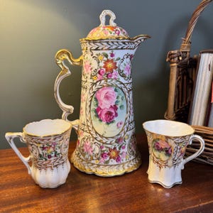 Könnte beinhalten: Eine Kaffeekanne aus Porzellan mit passendem Deckel und zwei Tassen, mit weißer Basis, verziert mit Goldrändern und bunten Blumenmustern, darunter rosa Rosen. Das Set zeigt kunstvolle goldene Henkel und gewellte Ränder an den Tassen.