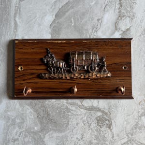 May include: A rectangular wooden wall-mounted key holder featuring a dark brown wood grain finish. A metallic copper-toned relief depicts a horse-drawn covered wagon with a driver. Three curved copper hooks are attached to the bottom edge.
