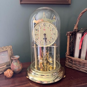 May include: A gold-toned anniversary clock with a glass dome, featuring a circular face with black Arabic numerals, floral accents, and the brand name Elgin. The clock has a rotating pendulum base with four gold pillars and black hands.