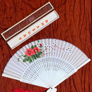 May include: A folding hand fan with a light beige fabric leaf featuring red camellia flowers and white plum blossoms. The fan has a white frame and a red tassel. It is accompanied by a rectangular box with the text 杭州王星記扇子.