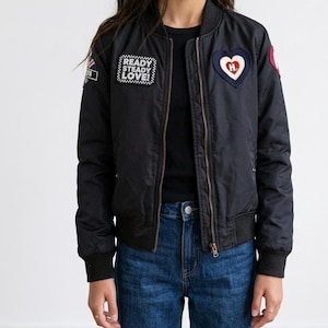 May include: A black bomber jacket featuring a front zipper, ribbed collar, and cuffs. The jacket is decorated with a rectangular patch reading READY STEADY LOVE, a heart-shaped patch with a red center, and a small flag patch on the right sleeve.