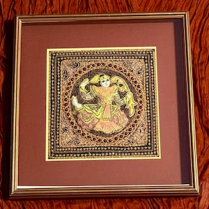 May include: Framed artwork featuring a central figure in a yellow and pink garment, surrounded by intricate embroidery in gold, brown, and black. The artwork is set against a burgundy mat and framed in gold, creating a decorative piece.